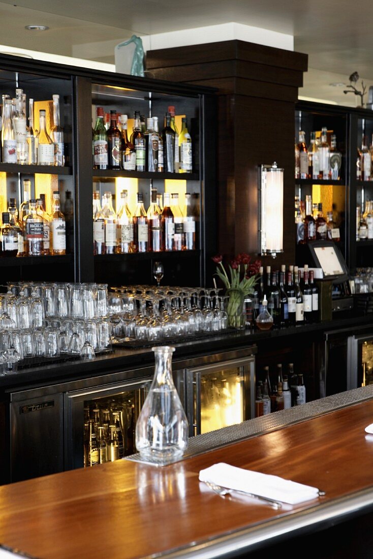 A polished wooden bar in a restaurant with shelves of spirits and glasses behind it