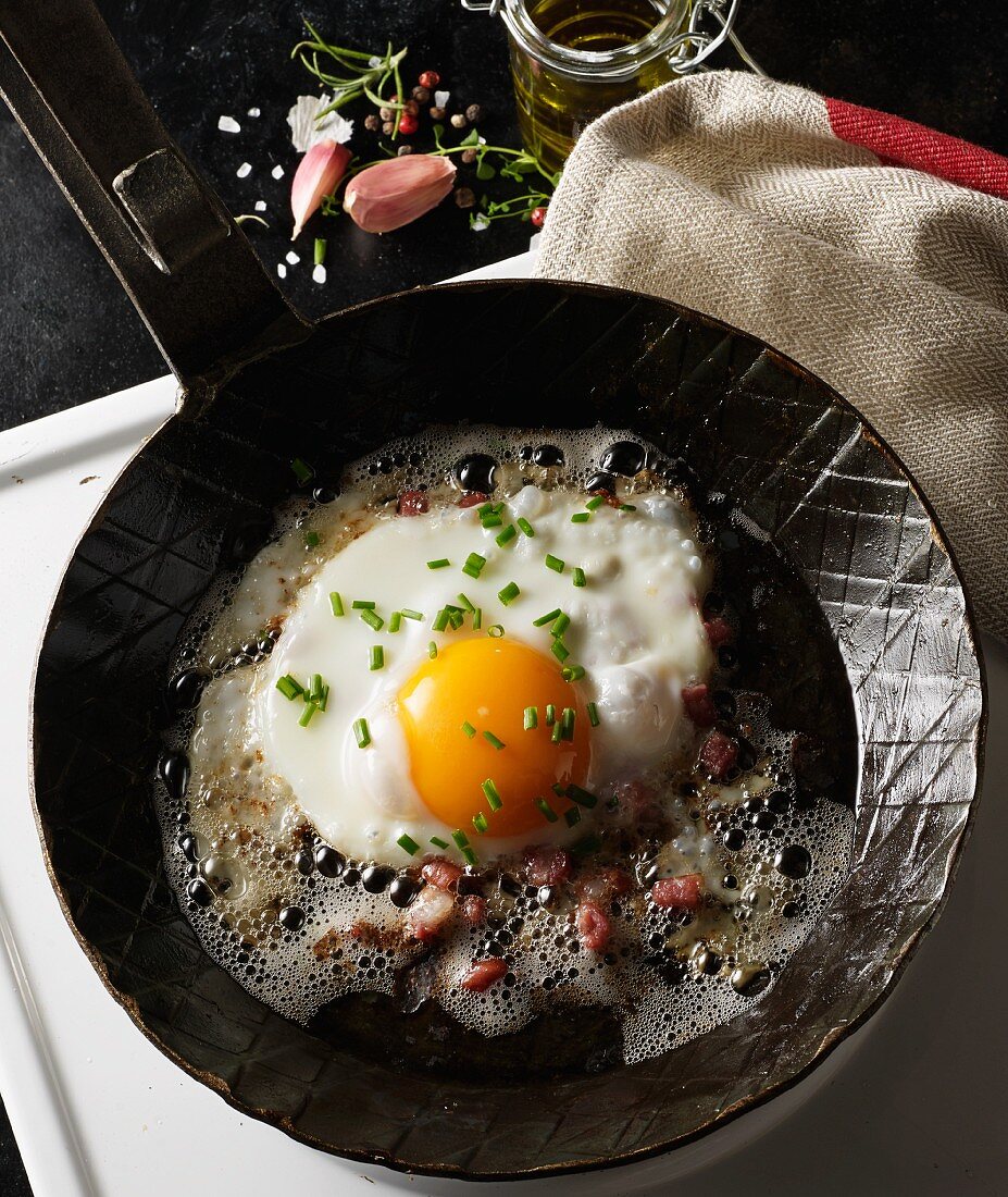 A fried egg with diced bacon and chives in a hot, black pan