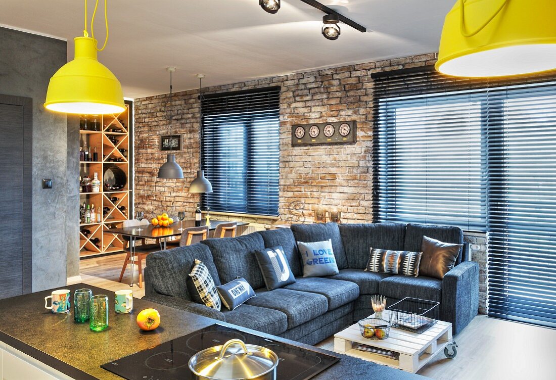 Grey sofa, dining area and wine rack in open-plan interior with brick wall and closed louvre blinds