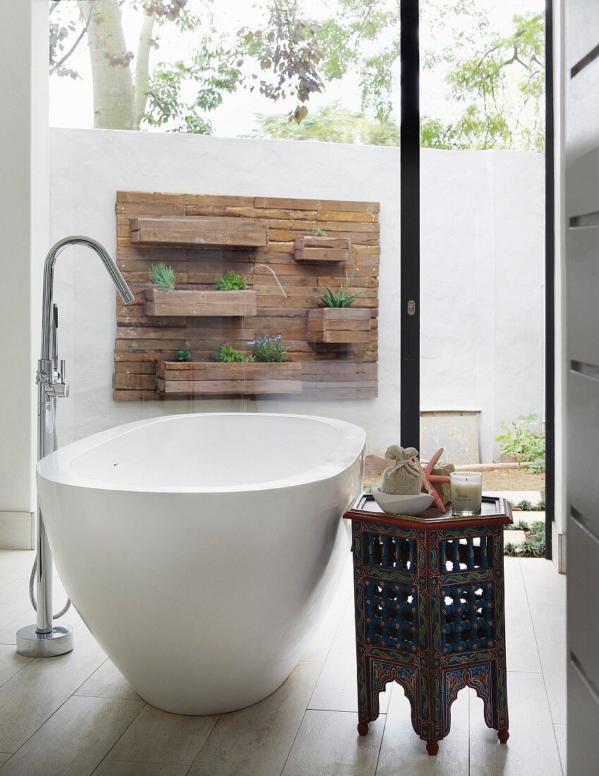Free-standing bathtub with standpipe tap fitting and Oriental side table in front of floor-to-ceiling window with view of patio