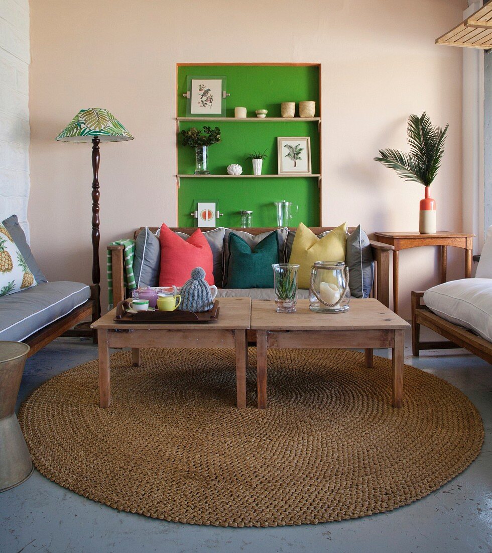 Wooden tables on round sisal rug in front of cushions on bench and ornaments in green-painted niche