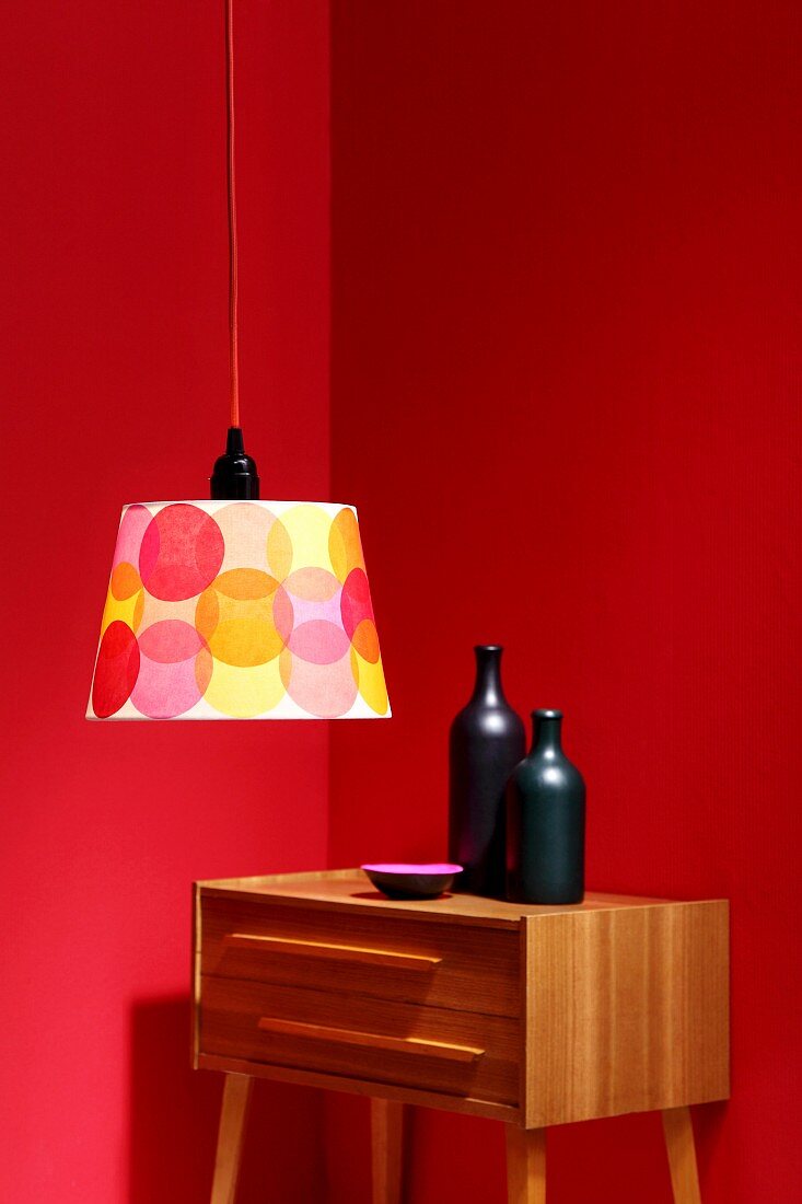 Simple lampshade decorated with colourful circles in front of ceramic bottles on retro chest of drawers against red walls