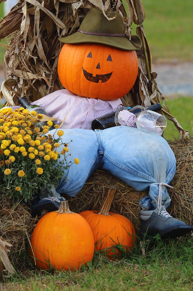 Pumpkin scarecrow wearing hat, shirt and trousers sitting on hay bale
