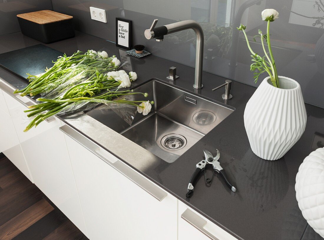 A designer kitchen counter with a stainless steel sink, a quartz stone work surface, a glass panel as a splash back, white buttercups and a white vase