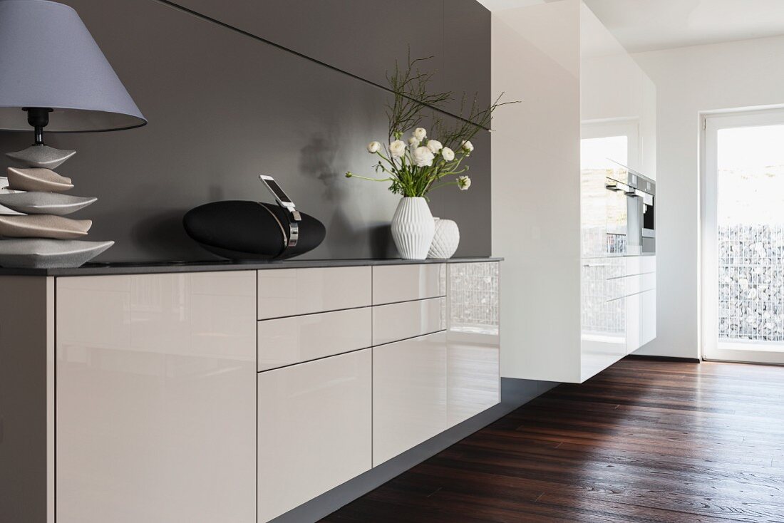 Weisses Designer-Sideboard, im Vordergrund Tischleuchte vor grauer Wand, im Hintergrund Kücheneinbauschrank mit Geräten