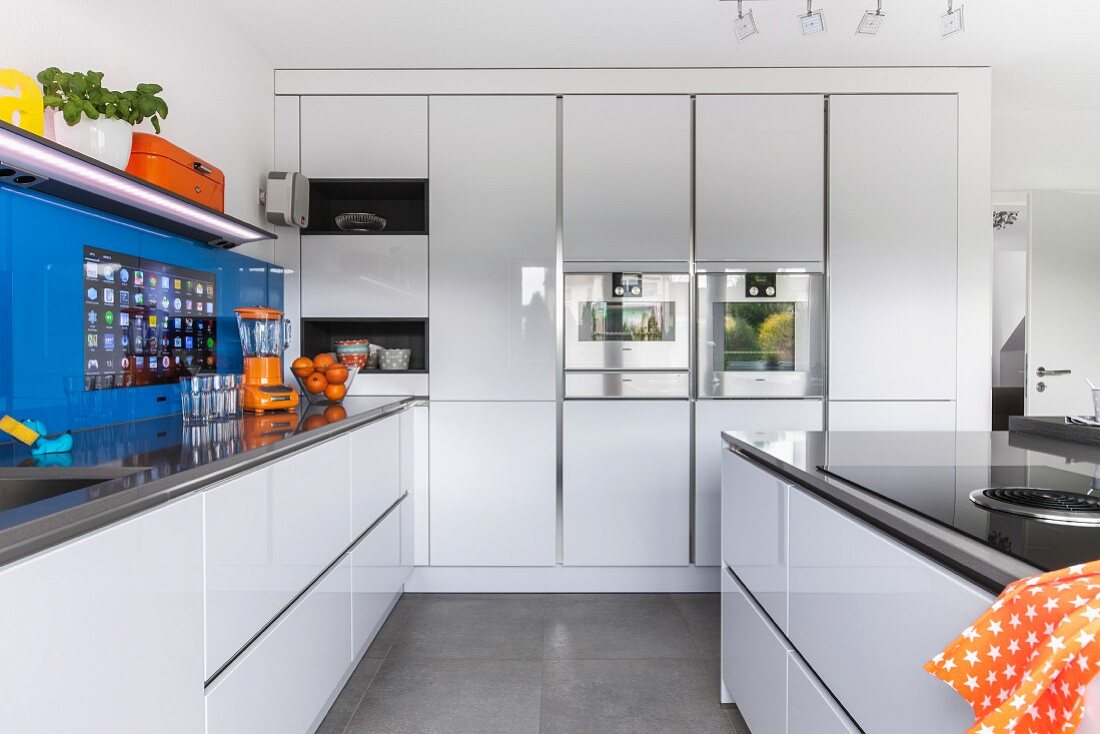 A white designer kitchen with a blue glass panel over the wall with an integrated touch screen to the side