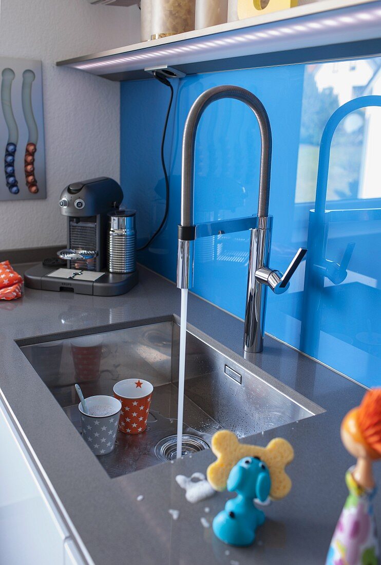 Water running into a stainless steel sink with a blue glass panel against the wall as a splash back