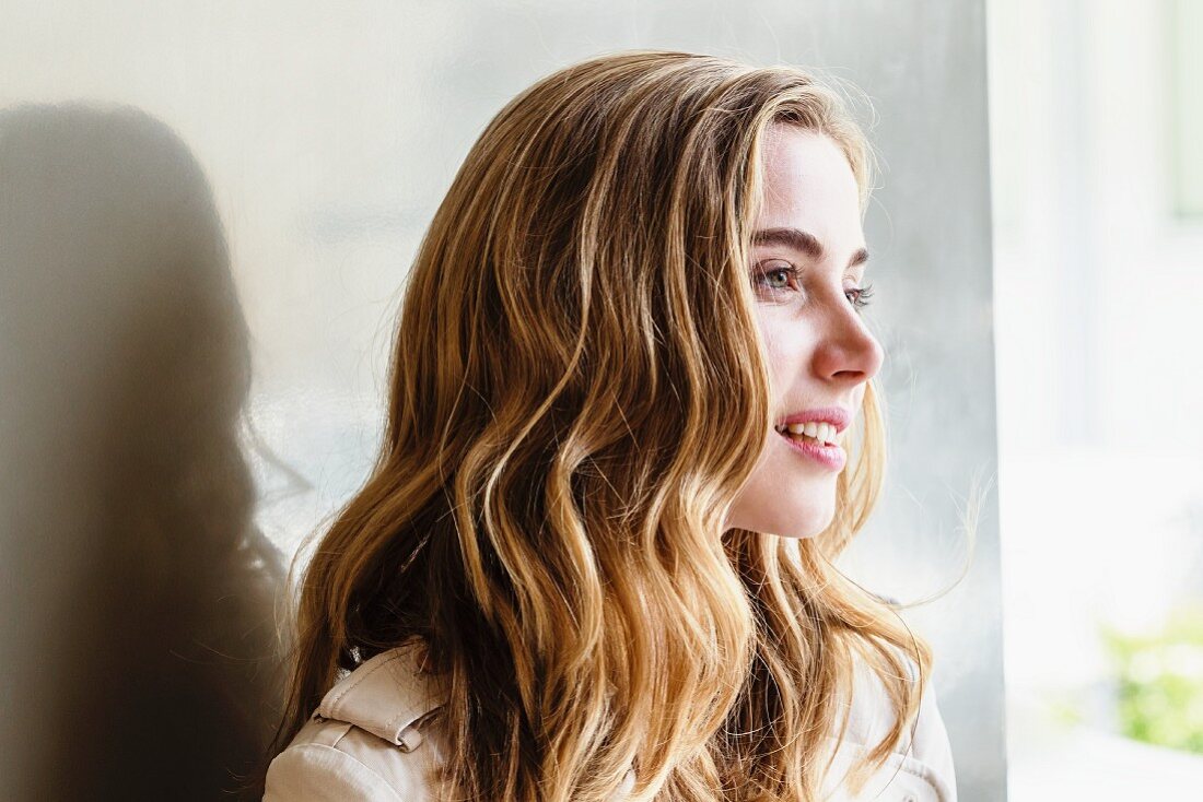 A young, blonde woman sitting by a window wearing a light jacket