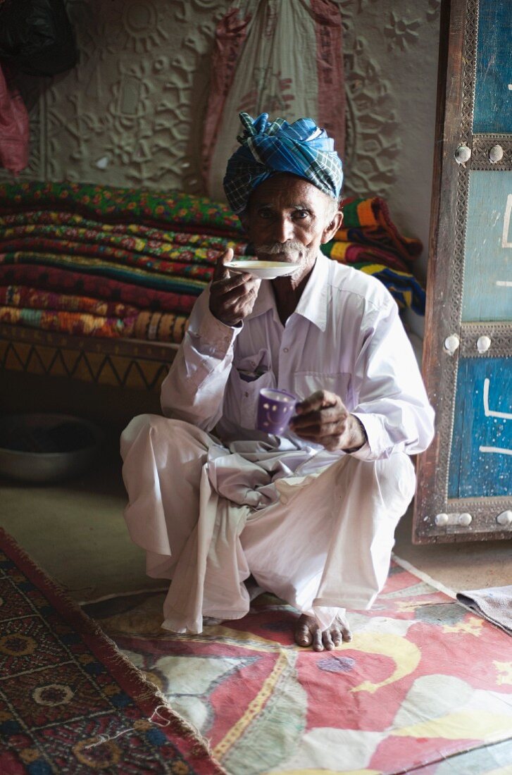Tee trinkender alter Mann mit Turban, vor einem Stapel handgeknüpfter Teppiche hockend, Soyla, Kachchh, Gujarat, Indien