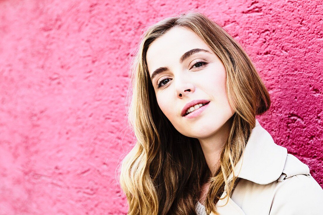 A portrait of a dark blonde woman against a pink wall