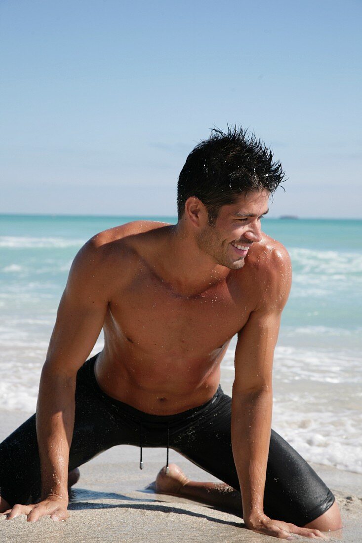 A young, topless man wearing bathing shorts keeling on a beach