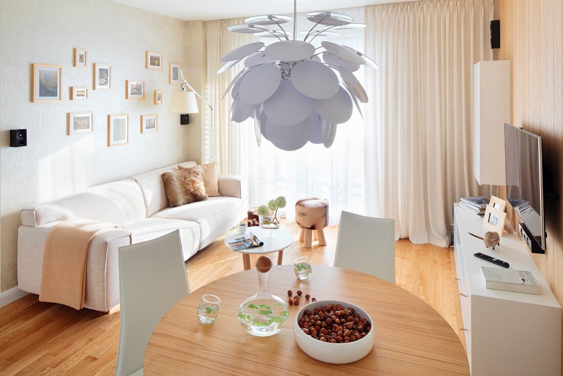 Designer pendant lamp with white leaves above round dining table and pale sofa in background in modern interior