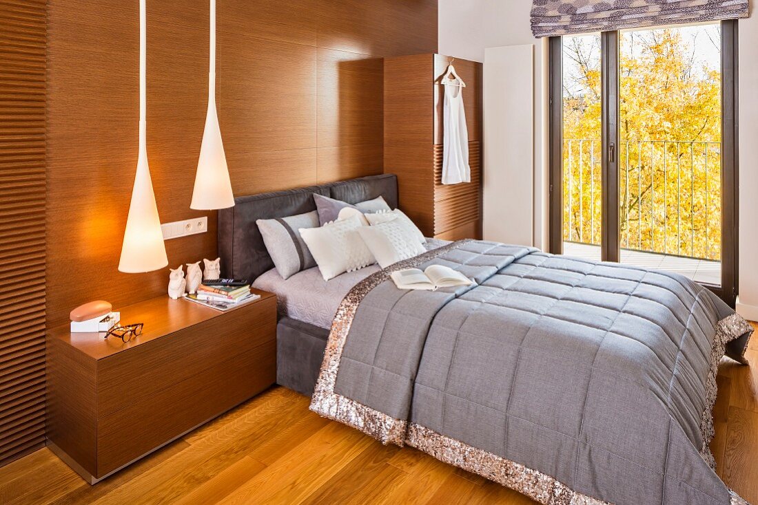 Double bed with grey bedspread against wood-clad wall with integrated cabinets