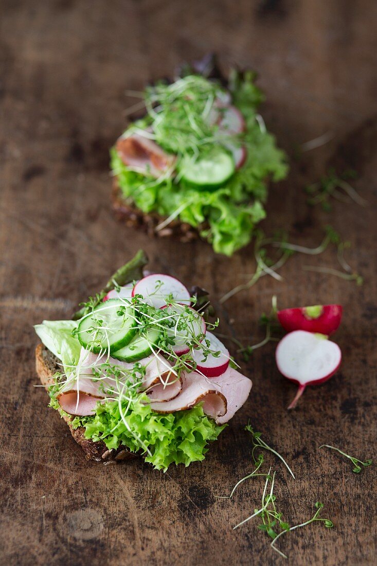Two open sandwiches with lettuce, ham, cucumber and radishes on a wooden surface