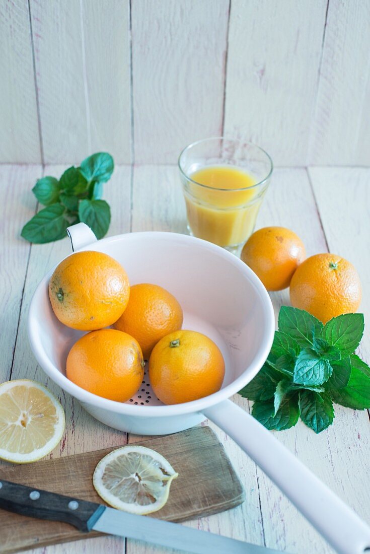 Orangen im Emaillesieb, frische Minze, Glas mit Orangensaft, Brett mit angeschnittener Zitrone