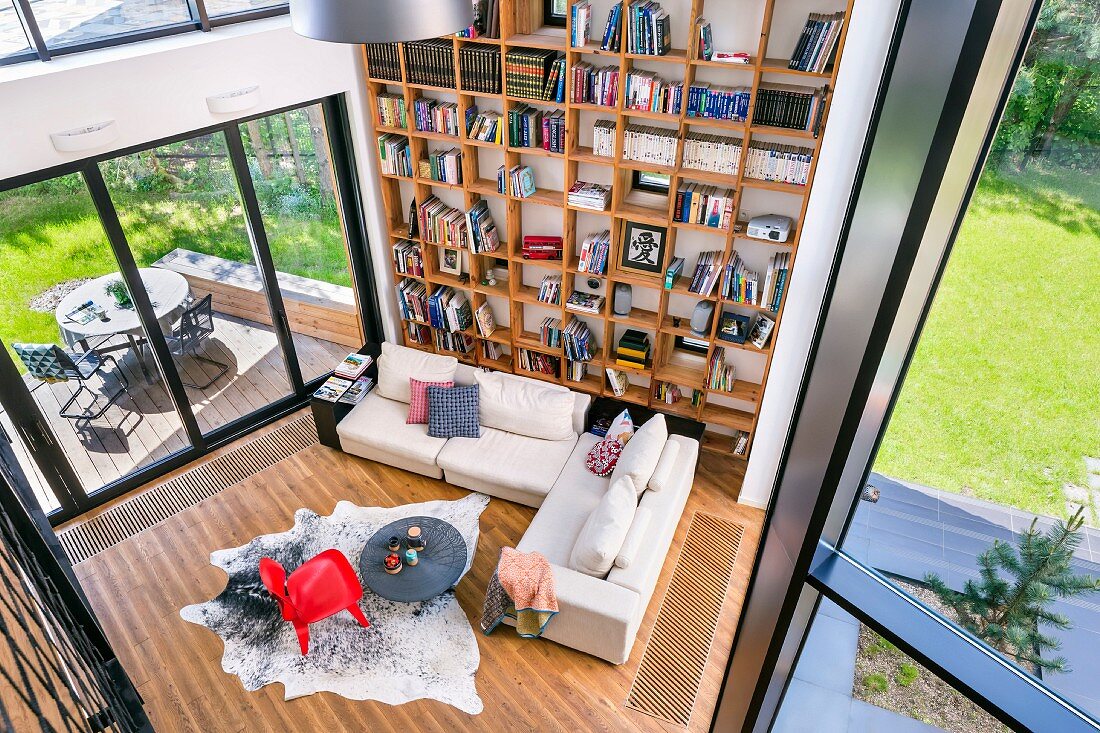 View down into living room with pale sofa set, red classic chair on animal-skin rug and tall wooden bookcase against wall