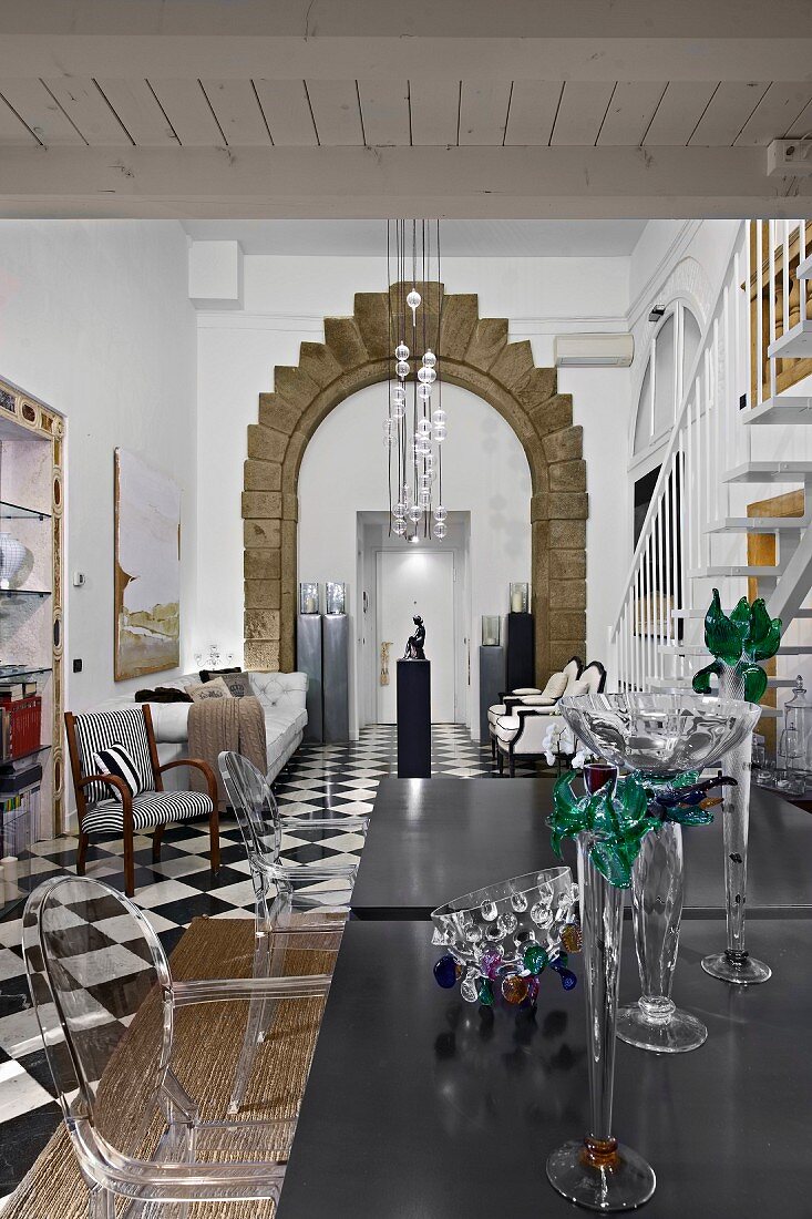 Glass vases on black table and Ghost chairs ni dining room with tall arched doorway with stone surround in background