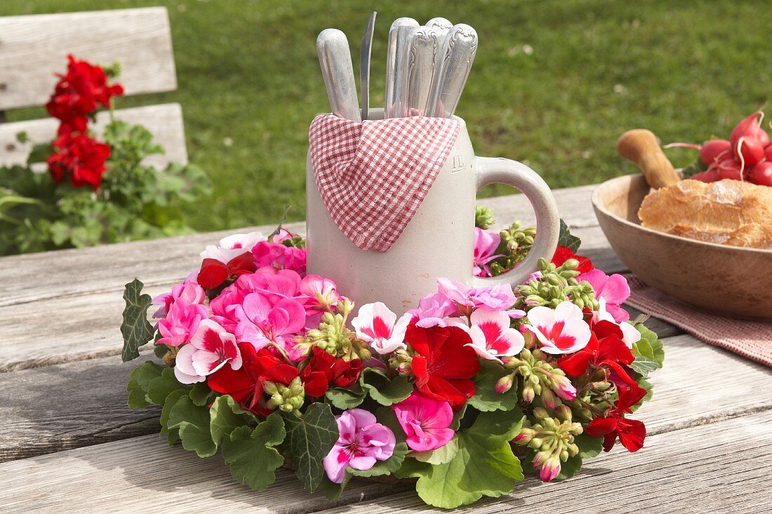 Cutlery in stein in wreath of geranium flowers on wooden table outdoors