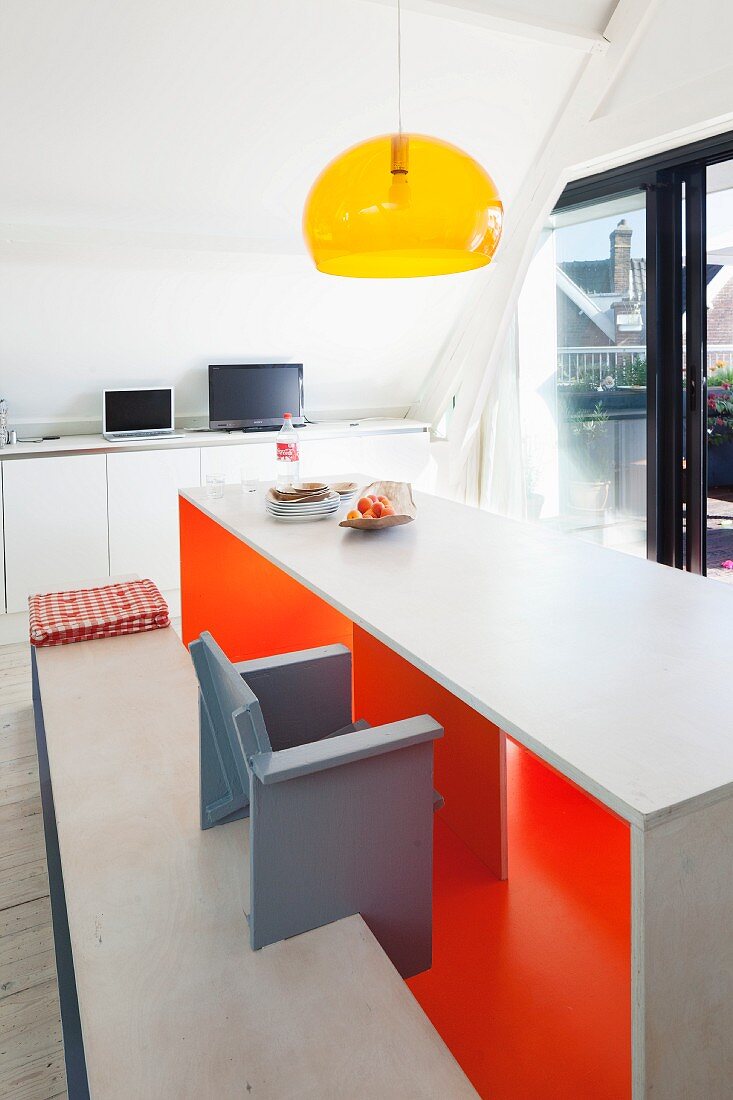 Designer table and bench set with brightly painted inner surfaces below pendant lamp with transparent yellow lampshade