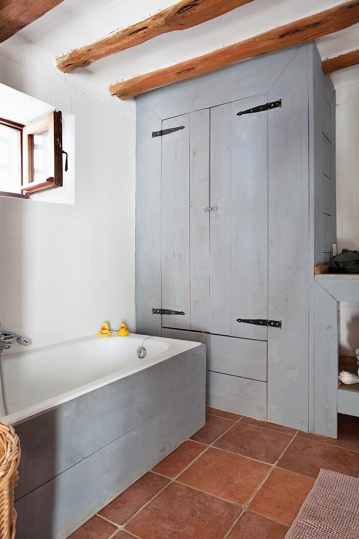 Badewanne mit hellgrau lasierter Holzfront und Einbauschrank in gleicher Optik in rustikalem Bad, mit Terrakottafliesen