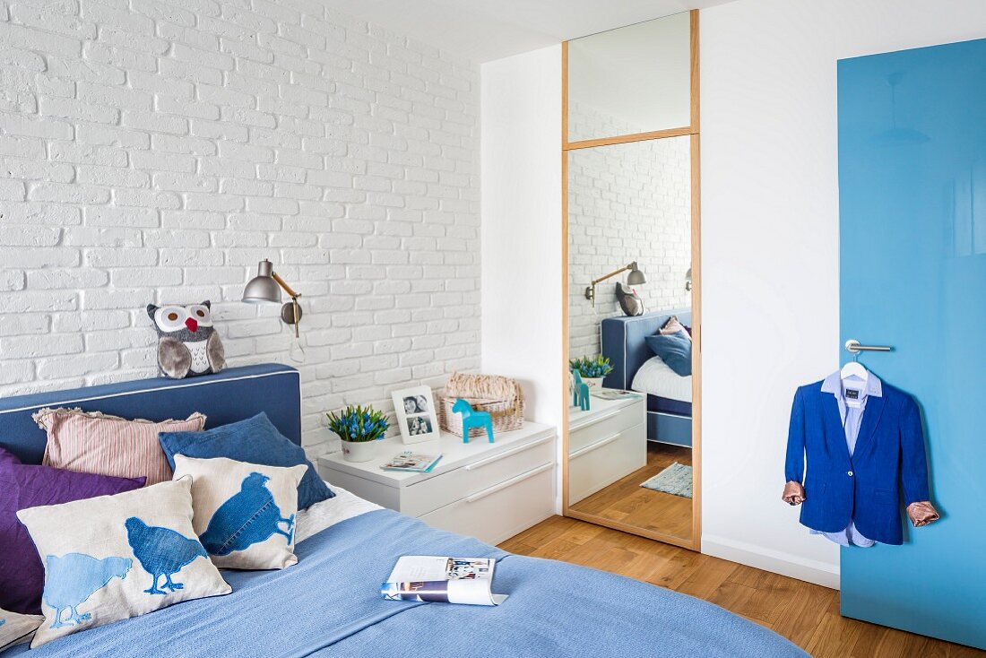 Double bed in shades of blue against whitewashed brick wall and full-length dressing mirror in bedroom