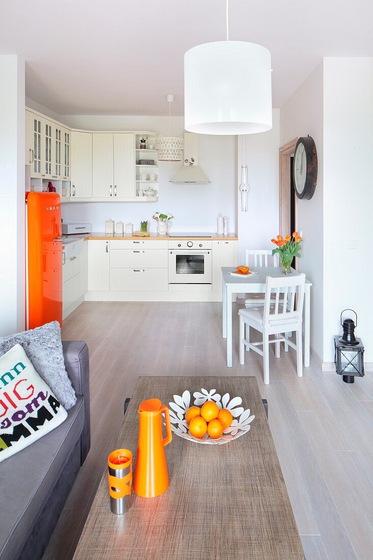 View across coffee table with orange accessories into open-plan kitchen with retro fridge and small dining area