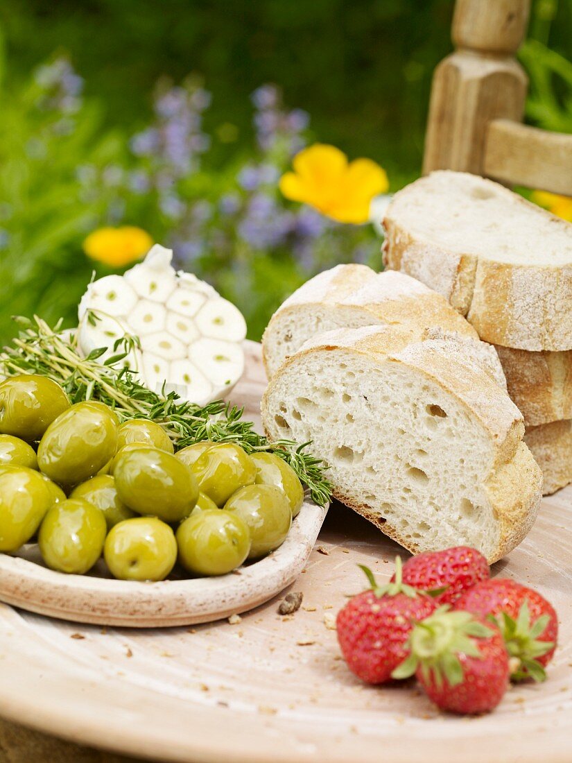 A sliced Milan baguette with olives, strawberries and garlic on a wooden board in the garden