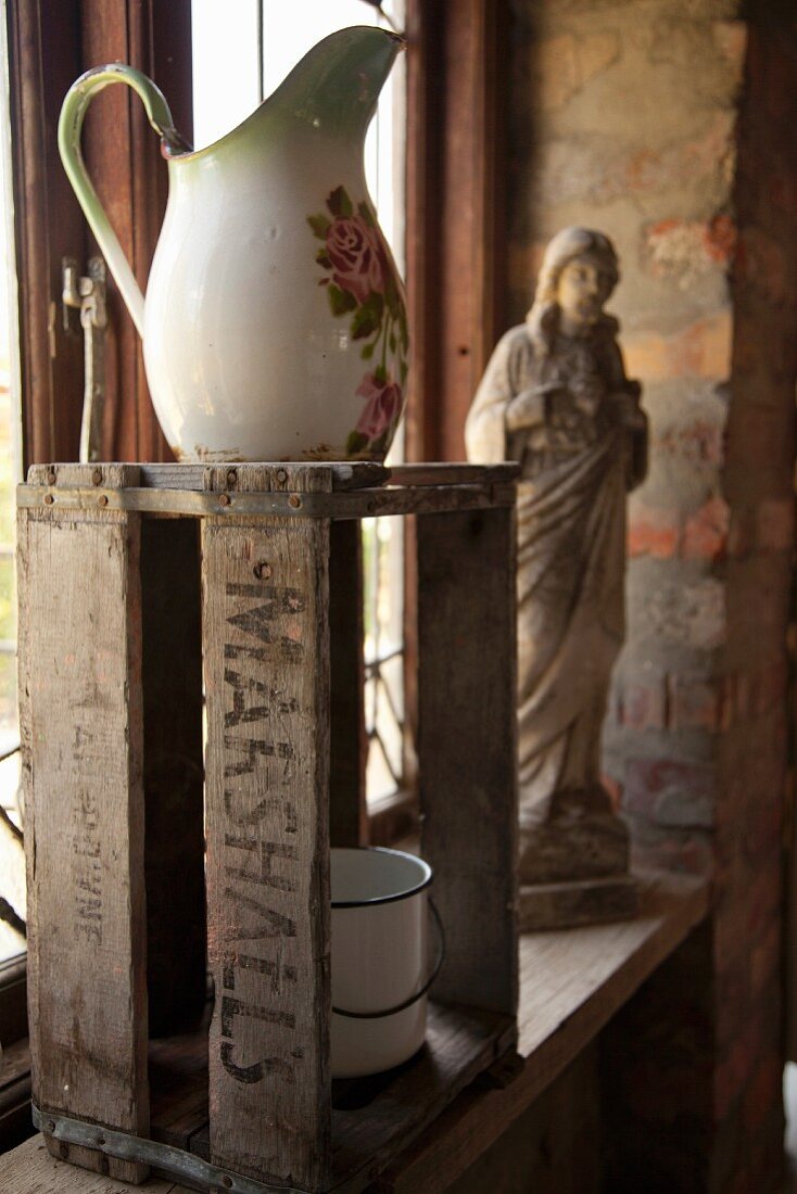 Old washstand pitcher on wooden crate in front of religious statue on windowsill