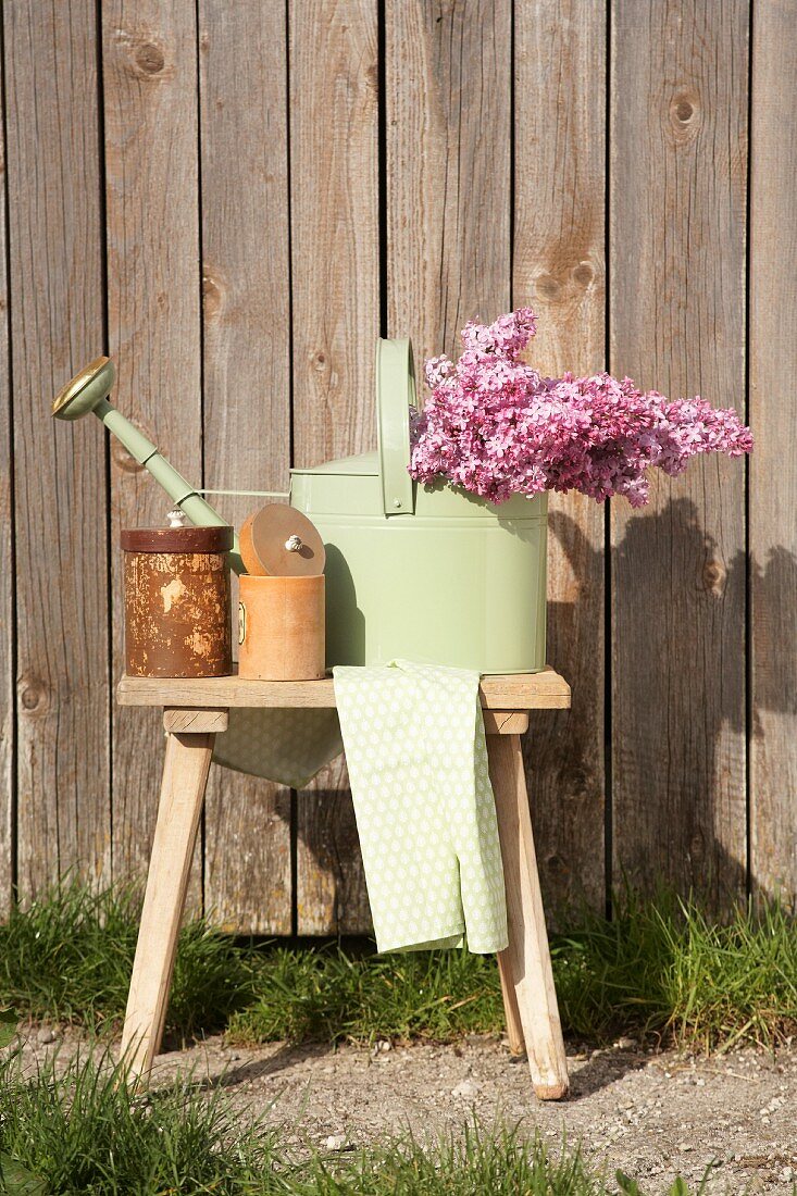 Bouquet of lilac in pastel green watering can and storage jars on wooden stool against wooden wall in outdoor sunshine