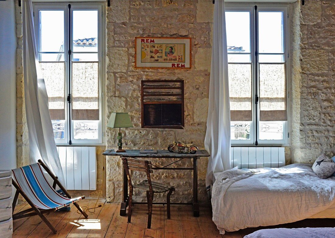 Mediterranean interior with desk and chair in front of rustic stone wall flanked by tall windows; single bed to one side