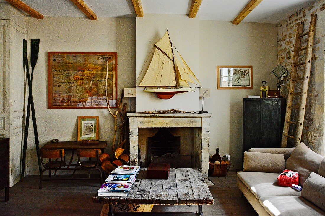 Rustikaler Couchtisch mit Platte aus Holzbrettern, im Hintergrund offener Kamin, darüber Modell Segelboot an Wand, in schlichtem Wohnzimmer