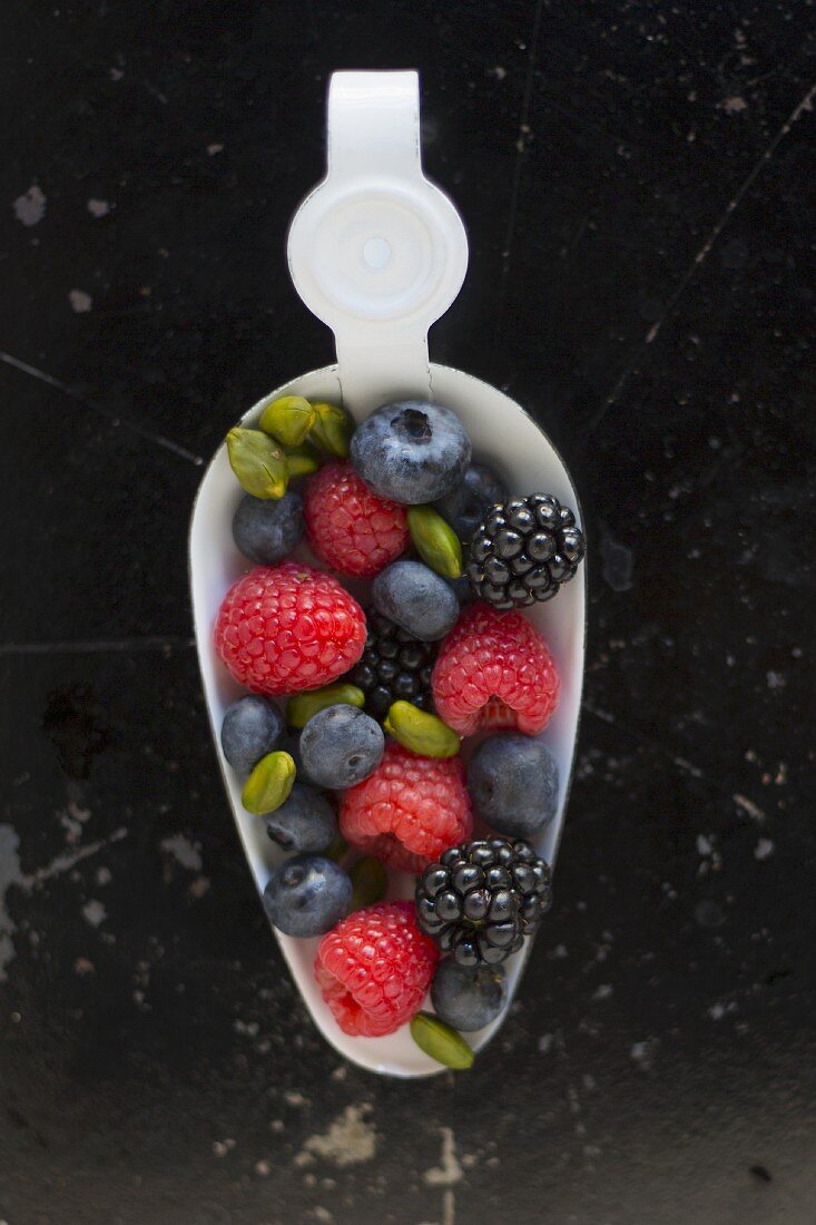 Blackberries, raspberries, blueberries and pistachios on a white enamel spoon