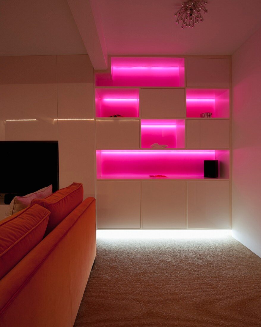 Pink beleuchtete Regalwand hinter einem orangefarbenem Sofa im dunklen Raum