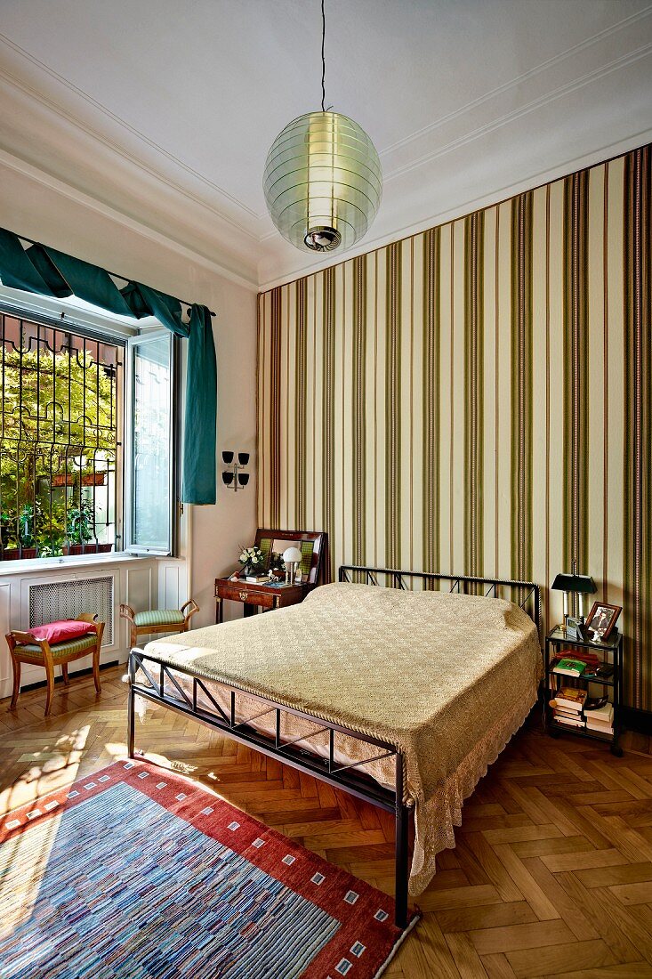 Double bed with modern, metal bedstead against wall with vertically striped wallpaper in bedroom with traditional ambiance