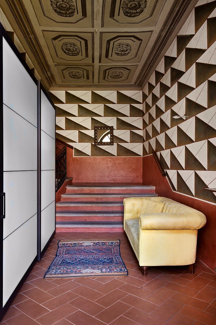 Modern, white cupboards with sliding doors and pastel yellow sofa at foot of staircase with terracotta red dado, expressive, graphic pattern on walls and old, coffered ceiling