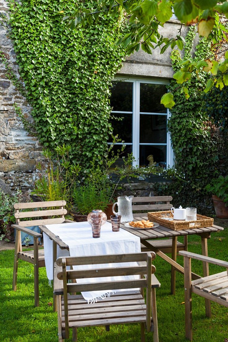Wooden table and chairs in garden outside stone house with ivy-covered façade