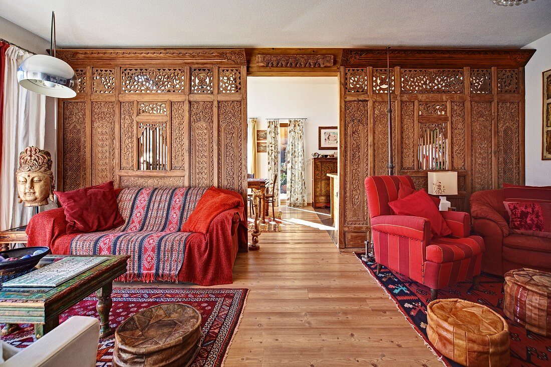 Kunsthandwerklicher Raumteiler in offenem elegantem Wohnraum; Sofa und Sessel in Rottönen, orientalische Lederpoufs mit Tisch und Ethnokunst - in einem Fertighaus