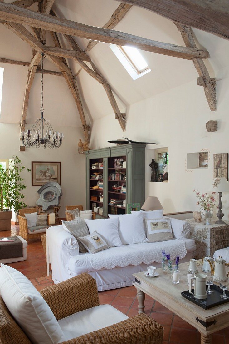 Exposed wooden roof structure above wicker armchairs and and white, loose-covered sofa in open-plan interior