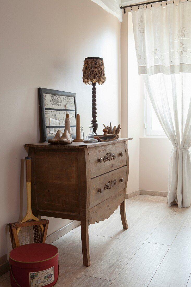 Wooden chest of drawers with tall legs next to draped lace curtain on window in simple, rustic bedroom