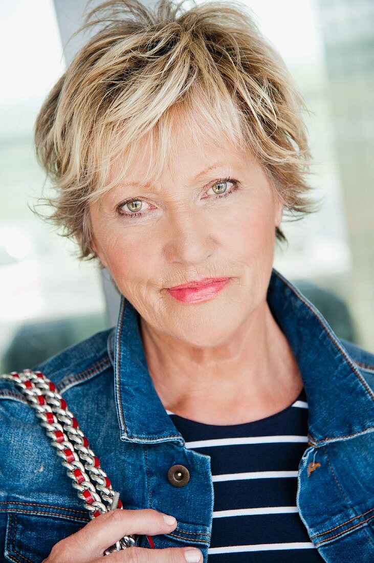 A portrait of an older woman in a denim jacket and a stripped shirt
