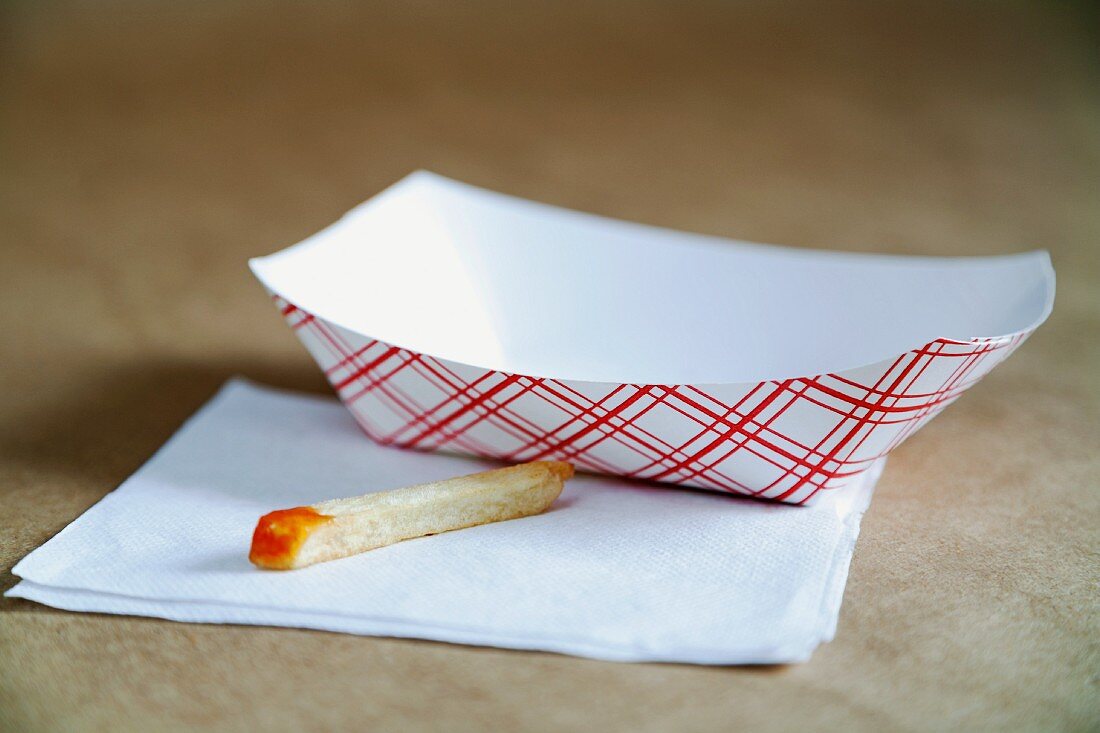 A single French fry with ketchup on a napkin with a paper dish