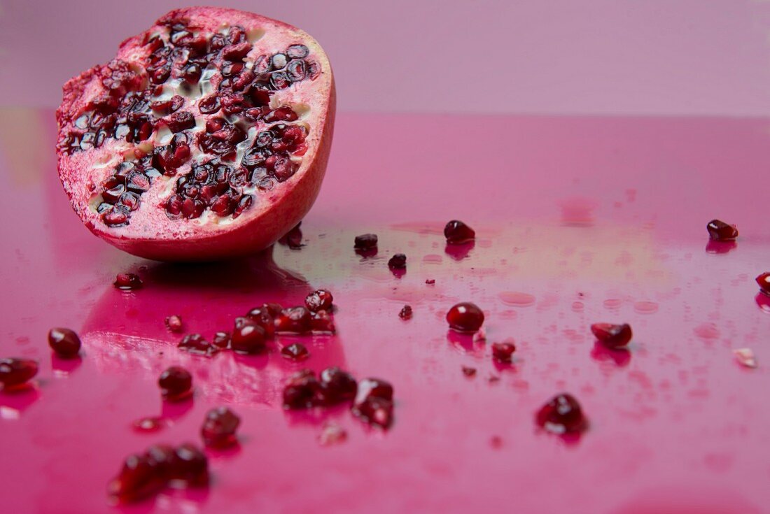 Half a pomegranate with seeds strewn around on a pink surface