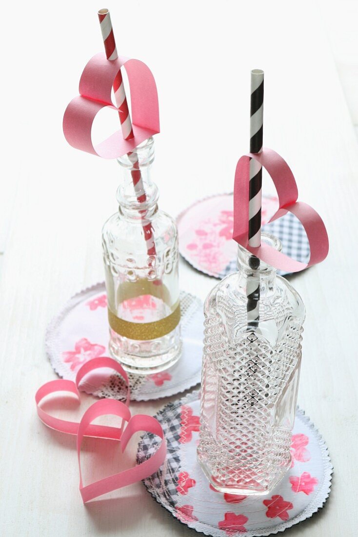 Two drinking straws decorated with pink, paper hearts in retro glass bottles on painted coasters