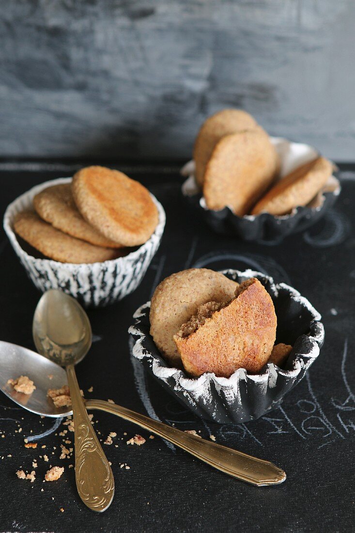 Kekse aus Mandelmehl und Honig in handgemachten Tonschälchen, Löffel mit Goldstiel auf schwarzem Untergrund