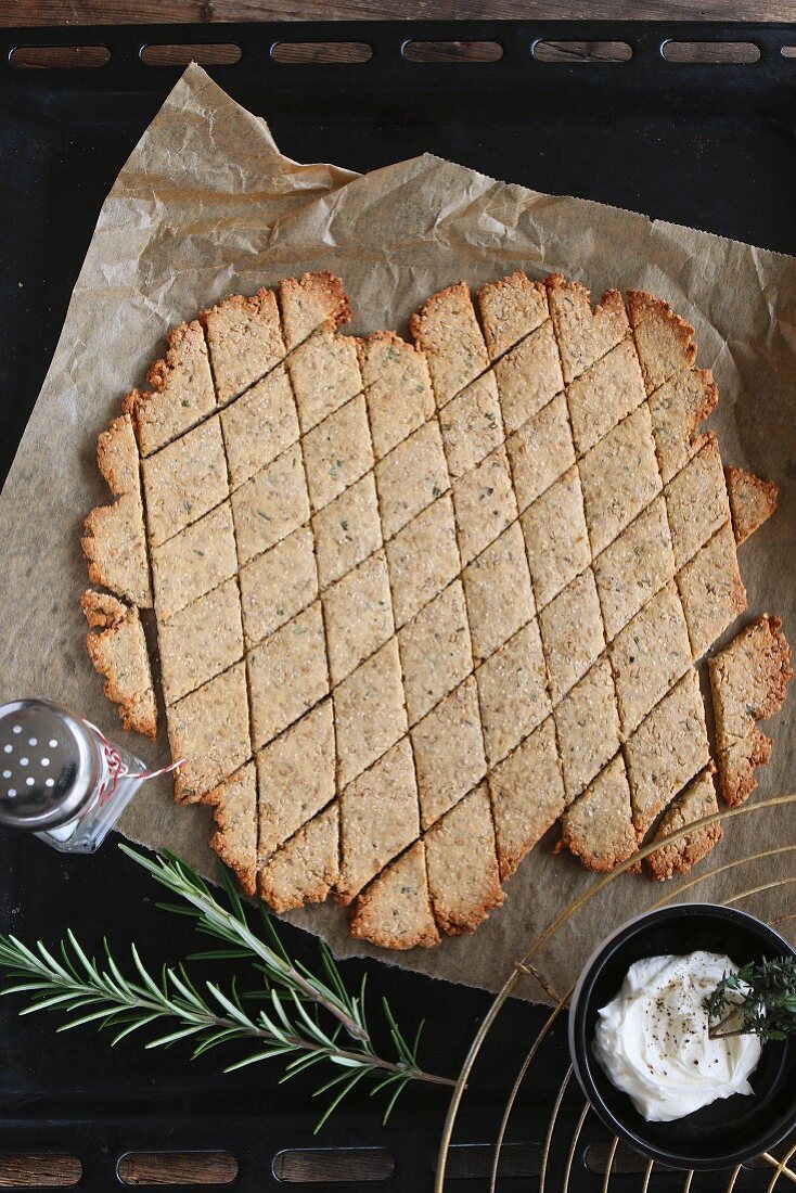 Gluten-free, diamond-shaped crackers with rosemary and sesame seeds on a baking tray with a bowl of dip and a sprig of rosemary