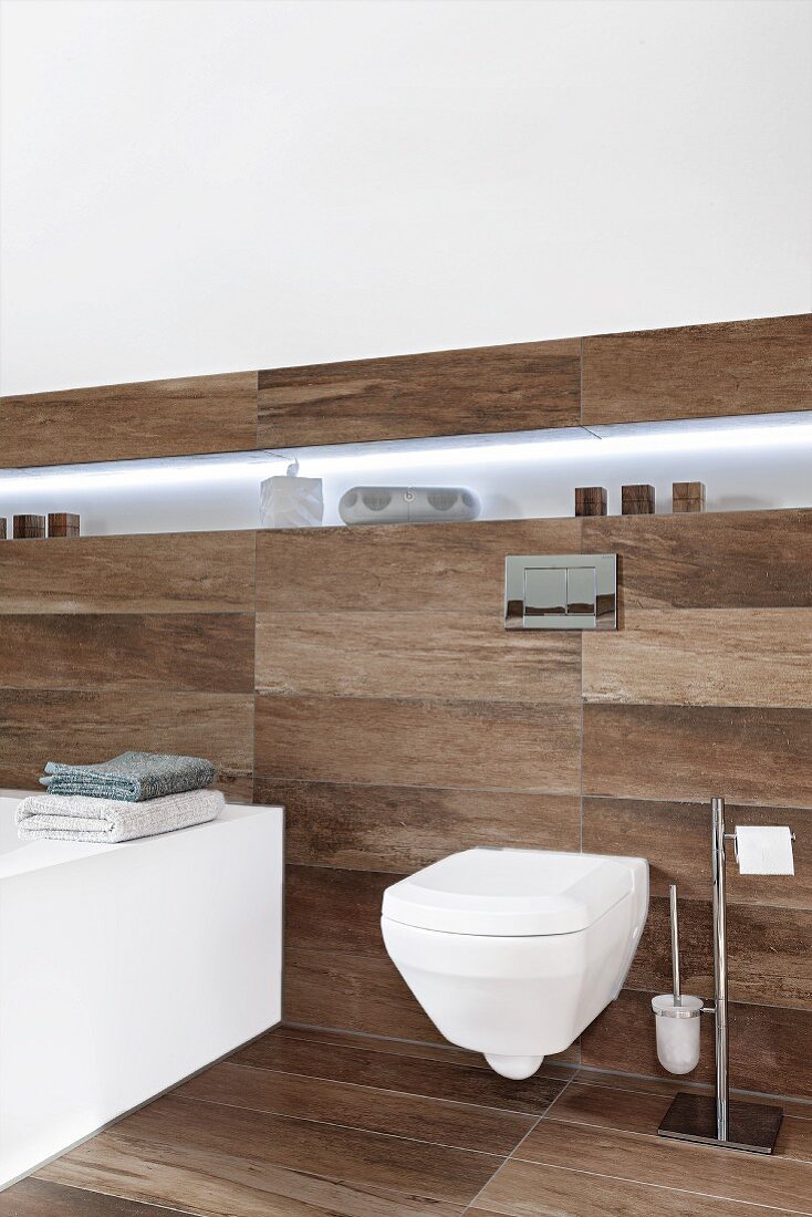 A modern, wall-mounted toilet and an illuminated shelf in a designer bathroom with dark, wooden-style tiles