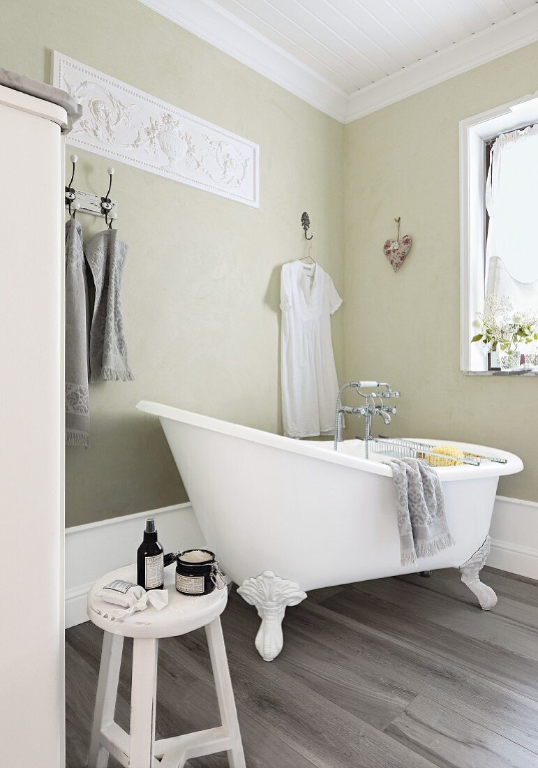 A romantic country house bathroom with a retro bathtub and a decorative relief on a pale green wall with grey, wooden-style floor tiles