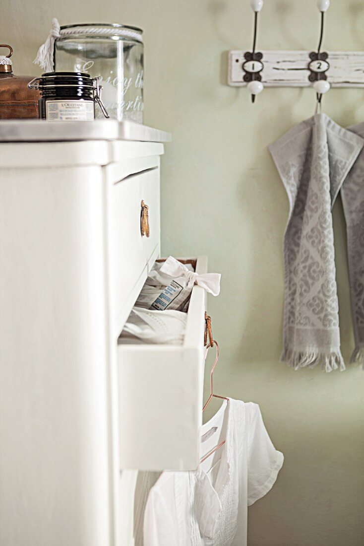 A white country house chest of drawers with a half-opened drawer and a row of vintage hooks on a pale green bathroom wall