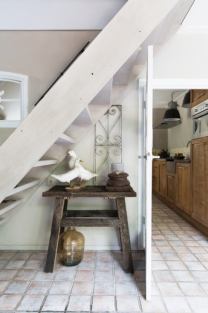 Rustic wooden table below white-painted wooden staircase in hallway with pale terracotta floor and view into kitchen through open door to one side