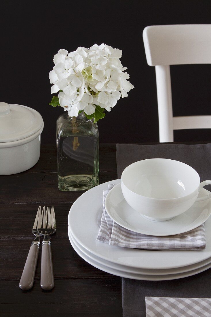 Cup and saucer on stacked plates in front of white hydrangea in vintage glass bottle and dark background